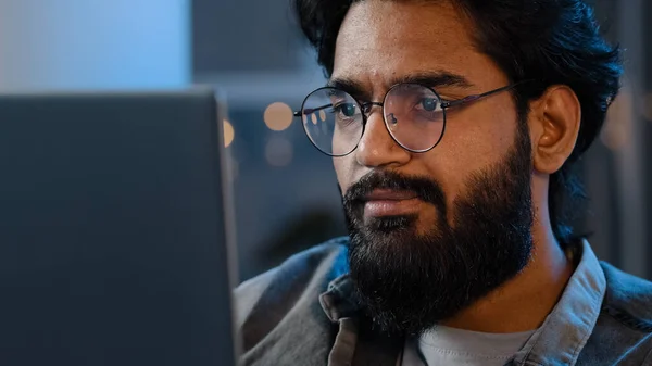 Close-up bearded male face reflection light from laptop monitor in glasses. Busy serious focused Arab Indian man looking at computer working freelance in evening playing game at night surfing in net —  Fotos de Stock