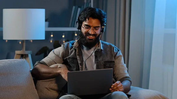 Árabe indiano engraçado homem barbudo feliz usa fones de ouvido sem fio se divertindo ouvindo música com laptop em casa sentado sofá noite tarde tempo escuridão quarto relaxante canto música desfrutar de áudio app — Fotografia de Stock