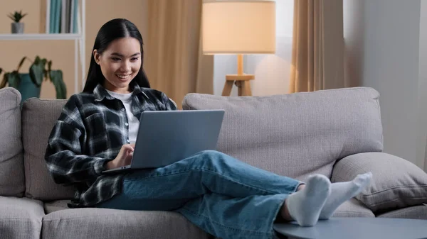 Smiling asian woman using laptop casual browsing searching online play computer game. Korean student girl freelancer sitting on couch e-learning internet education chatting studying working with wi-fi — Stockfoto
