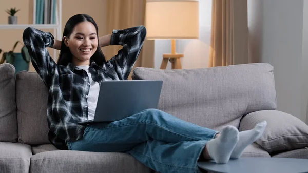 Asian woman Korean girl freelance student lady user sitting on sofa with hands behind head resting work break stop e-learning watching movie online video laughing enjoying leisure time with computer — Stok fotoğraf