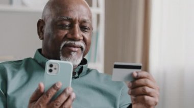Elderly African Dominican old mature man at home online transaction buying hold smartphone credit card successful payment buy on internet with mobile app food delivery order with phone easy e-payment
