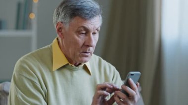 Mature elderly mature 60s caucasian man with phone male grandfather uses mobile app at home buys medicine online brows on net writes message to children in smartphone technology and old generation