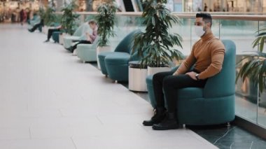 Sağlık maskeli genç Hintli adam salgın sırasında sağlığını koruyor alışveriş merkezindeki koltukta oturuyor dinleniyor sinirleri gevşiyor ve randevu için uzun bir bekleme konseptini bekliyor.