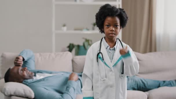 Malá africká americká dcera hrát s tátou otec pacient ležel na gauči dítě dívka předstírat, že je lékař při pohledu na kameru s úsměvem ukazující palec nahoru žádný problém vynikající zdravotní schválení gesto — Stock video