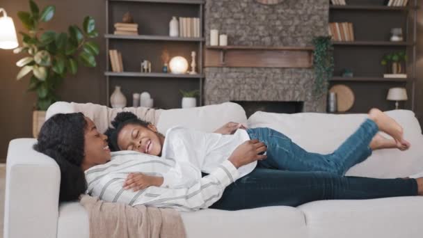 African mother with daughter cute beloved child girl lies on sofa at home in living room relaxing enjoying family bonding talking laughing hugging. Mommy and kid cuddling on couch comforting baby — Stock Video