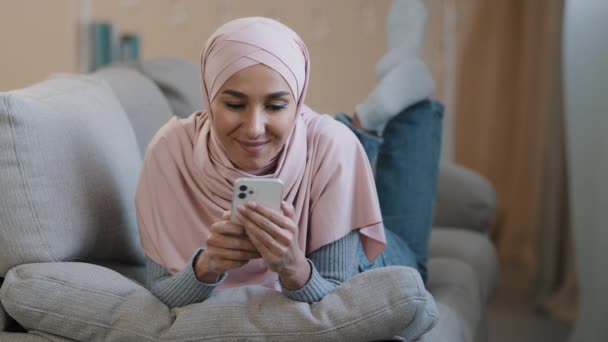 Genç, çekici, Arap müslüman kız İslamcı kadın kanepeye uzanmış mutlu Lübnanlı kadın tesettürlü giyiniyor cep telefonunda arkadaşlarıyla internetten komik video alışverişleri seyrediyor. — Stok video