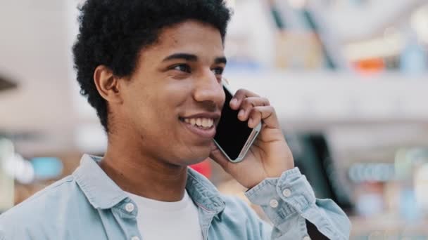 Африканський веселий привітний африканський чоловік із смартфоном у приміщенні розмовляє з друзями, які посміхаються. Бірюзовий хлопець має приємні розмови з мобільним телефоном. — стокове відео