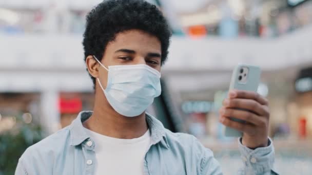 Портрет африканського домініканця, хворий чоловік, який має захисну медичну маску з телефоном, що показує жест в камері, рекомендує добре медичне застосування, задоволене онлайн-ліками, порядок бронювання послуги в смартфоні — стокове відео