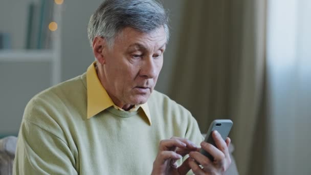 Зрелый пожилой человек 60-х годов белый мужчина с телефоном дедушка использует мобильное приложение дома покупает лекарства онлайн брови в сети пишет сообщение детям в технологии смартфона и старого поколения — стоковое видео