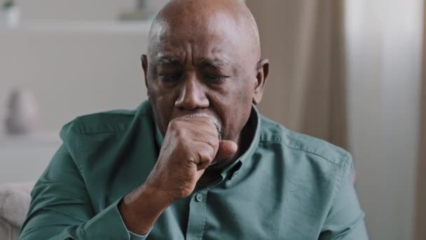 Афроамериканец нездоровый седой старик 60-х годов, кашляющий с симптомами гриппа, сидит на диване дома больной взрослый дедушка с болью в горле, коронавирусная инфекция — стоковое видео