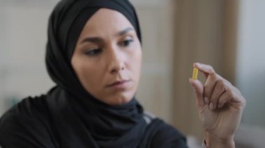 Young female thoughtful patient arabian islamic girl in hijab hold yellow medicine capsule vitamin tablet in hand lebanese doubtful sick woman look on healthy drug pill in front health care concept