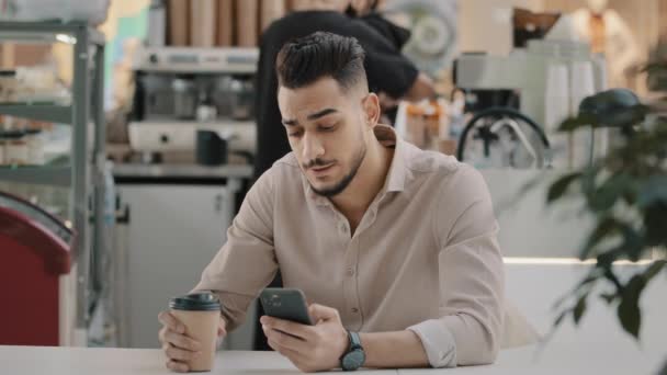 Indian hispanic arabian man multiracial businessman sitting at cafe table drinking hot coffee tea from paper cup looking in mobile phone typing smartphone chatting browsing using online app free wi-fi — Video Stock