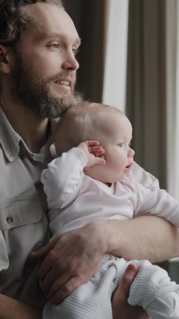 Pionowy widok kaukaski dorosły ojciec kochający opiekuńczy tatuś trzymający dziecko córka mały syn noworodek w rękach patrząc przez okno stojący w domu rano dotyka dziecka ręka rodzicielstwo więź — Wideo stockowe