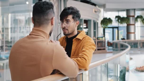 Twee jonge positieve Indiase jongens staan in de lobby te praten over zakelijke probleem zorgeloze vrienden bespreken delen nieuws vriendschap genieten weekend in vrije tijd Arabisch man drinken sap terug uitzicht — Stockvideo