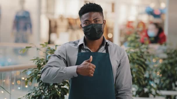 Jonge zelfverzekerde Afrikaans-Amerikaanse man in medische masker ober in schort winkel assistent cafe eigenaar op zoek naar camera poseren duim omhoog gebaar van goedkeuring reclame catering inrichting — Stockvideo