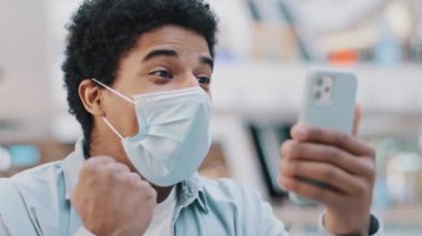 Portrait african american man guy wearing medical mask with phone wins online bonus mobile game gets financial victory receive good message goal makes victory hand gesture triumph smartphone success