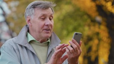 Mutlu yaşlı, gülümseyen, olgun beyaz erkek büyükbaba cep telefonuyla uzaktan video görüşmesi yapıyor. Online sohbet olumlu yaşlı adam aile içi telefon kullanımıyla modern kablosuz teknoloji kullanıyor.