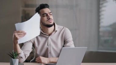 Hispanic exhausted male business man annoyed guy suffer from summer heat in office home without air conditioner feeling hot high air temperature uncomfortable waving papers cooling make wind freshen