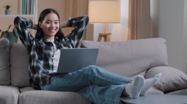 Asian woman Korean girl freelance student lady user sitting on sofa with hands behind head resting work break stop e-learning watching movie online video laughing enjoying leisure time with computer
