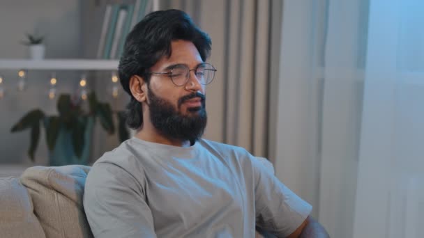 Задумчивый арабский задумчивый индийский взрослый бородатый мужчина сидит дома на диване и смотрит в сторону, думая, улыбаясь в камеру. Мужчина владелец собственной квартиры арендатор наслаждается отдыхом на диване — стоковое видео