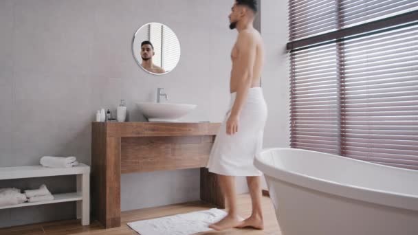 Naked bare sexy muscular Arab Indian man enters going walking in bath after shower with white towel on hips washes face with warm water in bathroom sink looks at reflection in mirror morning hygiene — Stock Video