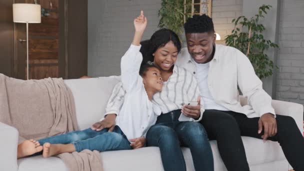 Αφρικανική οικογένεια γονείς με παιδί κόρη κορίτσι χαλαρώστε στον καναπέ σπίτι σαλόνι χρησιμοποιώντας εφαρμογές smartphone επιλέγοντας στο internet κατάστημα κράτηση τροφίμων παράδοση με κινητό τηλέφωνο μαζί κοιτάζοντας την οθόνη — Αρχείο Βίντεο