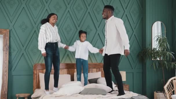 Mutlu Afro-Amerikan ailesi, genç anne-babası ve beyaz kız çocuğu, sabah evde el ele tutuşuyor, yatakta zıplıyor, gülüyor, yatak odasında komik oyunlar oynuyor. — Stok video