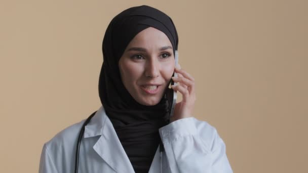 Arabian musulmán joven cirujano practicante usar médico abrigo tradicional hijab hablar por teléfono celular consultar paciente remoto mujer islámico terapeuta discutir tratamiento móvil conversación en clínica — Vídeo de stock