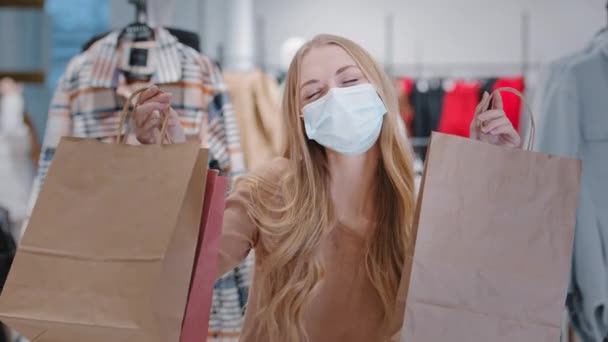 Πλούσια ευτυχισμένη ξανθιά γυναίκα αγοραστής φορώντας ιατρική πρόσωπο προστατευτική μάσκα πελάτη κορίτσι στην μπουτίκ ρούχων με τσάντες ψώνια χορό απολαμβάνοντας μαύρο εκπτώσεις Παρασκευή επιτυχής αγορά ύφασμα πολυτελείας — Αρχείο Βίντεο