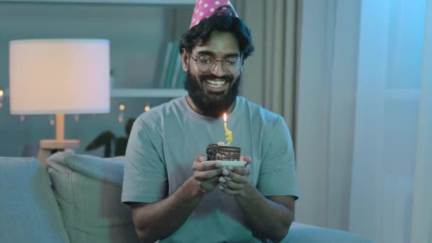 Otthon party boldog arab indiai szakállas férfi szemüvegben kalap tartja születésnapi torta szelet gyertya kívánság mondván beszél a jövő álom fúj ünneplés egyedül ül a kanapén — Stock videók