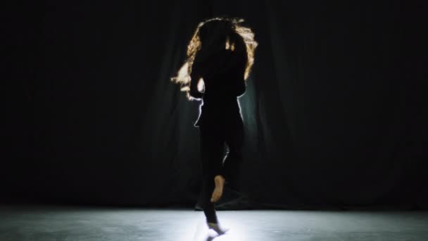 Aktive wunderschöne anmutige Mädchen mit langen lockigen Haaren in schwarzen Kleidern in dunklen Tanzstudio auf der Bühne in helle weibliche Silhouette Tänzerin Künstlerin Tanzbewegungen rhythmische zeitgenössische Bewegung zur Musik — Stockvideo