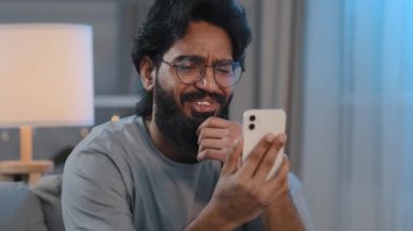 Arap sakallı, gözlüklü, cep telefonlu, video konferans konferans mesafesinden konuşan 30 'lu yaşlarda bir adam internetteki arkadaşlarıyla sanal iletişim kurmak için akıllı telefon kullanıyor.