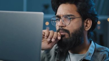 Odaklanmış Arap Hintli sakallı adam gözlüklü, gece geç saatlerde evde bilgisayar başında çalışan, ekran arama kararına bakan, problem çözümü üzerine düşünen online bir proje oluşturma şüphesi hissediyor.