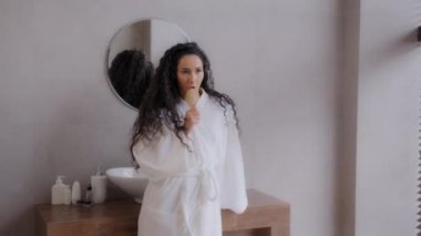 Komik, neşeli, bornozlu, genç, kıvırcık kadın banyoda en sevdiği müzikle dans ediyor. Saç fırçasını mikrofon gibi kullanıyor.