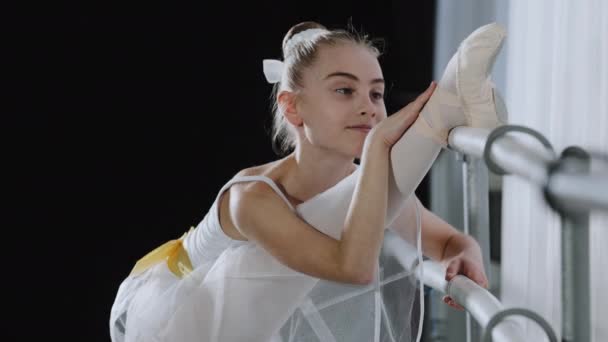 Bale etekli esnek balerin kız çocuğu dans sınıfında dans sınıfının yakınına uzanıyor. Bacaklarını zevkle esnetiyor. Kolayca esneklik alıştırmaları yapıyor. — Stok video