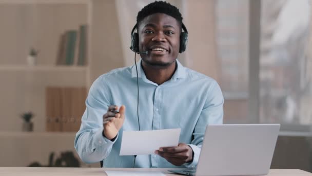 Jeune homme d'affaires enseignant afro-américain homme d'affaires porte un microphone tête enseignant étudiant en ligne en regardant la caméra fournir un soutien professionnel clients instruire à distance utiliser des documents financiers — Video