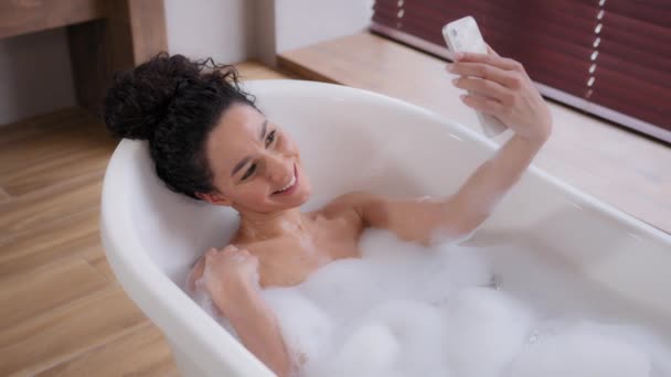 Genç mutlu İspanyol kadın blogcu. Lüks köpük banyosunda rahatlarken telefonla fotoğraf çekiliyor. Selfie pozu veriyor. — Stok video