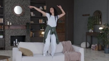 Genç, neşeli, neşeli, neşeli kadın ev hanımı modern oturma odasındaki kanepede aktif olarak dans ediyor ritmik müzik dinliyor boş zamanlarında stres olmadan tek başına hafta sonunun tadını çıkarıyor.