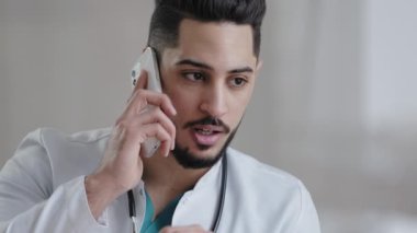 Ciddi erkek İspanyol doktor doktor doktor asistanı hasta danışmanı cep telefonu Arap danışman cerrahı uzaktan danışmanlık hizmeti veren klinik kabinesinde cep telefonu kullanıyor.