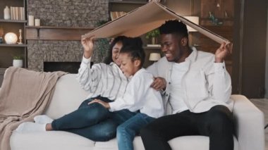Karton çatının altında koltukta oturan Afrikalı çift kameraya gülümseyip mutlu bir kızın koşuşuna bakıyor ve koltukta oturuyor. Yeni eve taşınma günü. Kendi ev sahibi portresine taşınma.