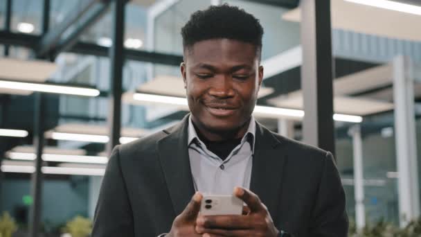 Χαμογελώντας χαρούμενος αφρικανός άνθρωπος διευθυντής γραφείου αφεντικό εργαζόμενος Αμερικανός άντρας επιχειρηματίας κατέχει smartphone scroll οθόνη κινητής συνομιλίας σε μέσα κοινωνικής δικτύωσης μήνυμα κειμένου app διαβάσει καλά νέα κάνουν τη σειρά στο διαδίκτυο — Αρχείο Βίντεο
