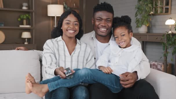 Carefree african american father, mather relaxing with biracial daughter child girl at hme holidays sitting on sofa at home watching television together. Cheerful family relaxing watching tv movie — Vídeo de stock
