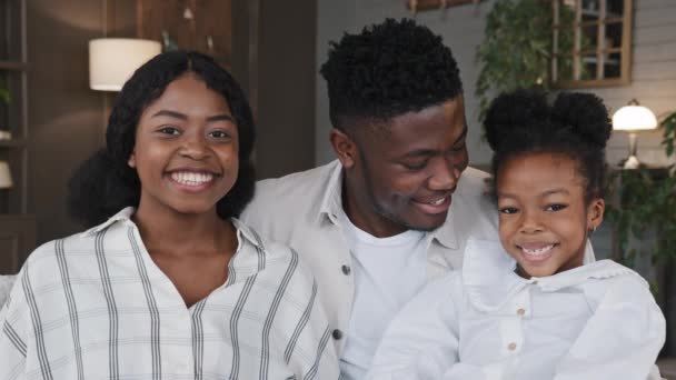 Heureux portrait de famille africain. Parents avec fille gaie fille regardant la caméra riant câlins à la maison souriant dents. Mère positive, père et enfant étreignant sur le canapé. Concept de santé dentaire — Video