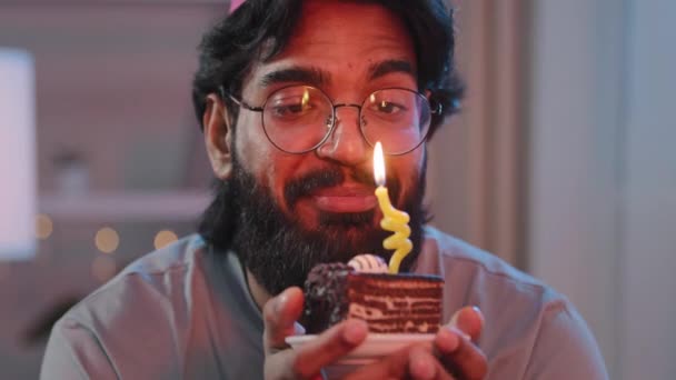 Retrato de hombre multirracial con gruesa barba negra en gafas y sombrero festivo con pastel en luz por la noche en el interior en la oscuridad dice deseo sueño felizmente celebra cumpleaños soplando vela ardiente — Vídeos de Stock