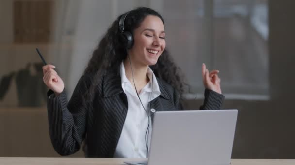 幸せな若いアラビア人の女の子の女性気楽な学生の労働者は、ヘッドフォンを着用音楽ダンス精力的な動きは、コンピュータの仕事が自宅のオフィスデスクで楽しい座っている後にリラックスした高品質の音の歌を楽しむ — ストック動画