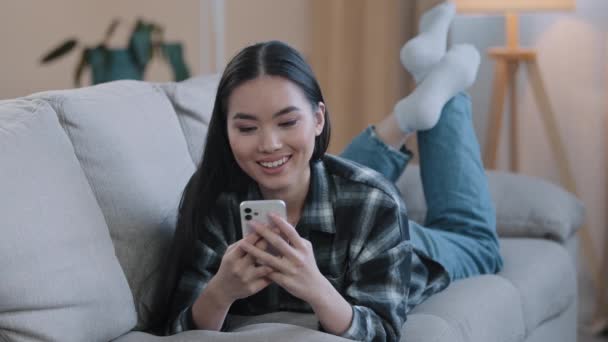 Ásia feminino relaxante menina mulher deitado no sofá em casa desfrutar de bate-papo digitando aplicativo celular relaxar assistir viral ao vivo vídeo jogar jogo siga como post em mídias sociais compartilhar e enviar sorrir emoji rolagem web — Vídeo de Stock