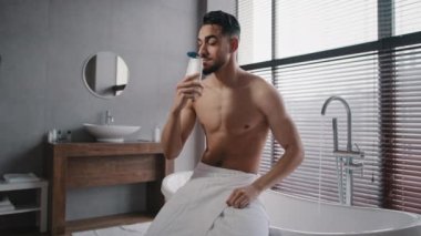 Arap Hintli, kaslı, çıplak, seksi, traşsız adam kalçasına beyaz banyo havlusu takıyor banyoda banyo yapmaya hazırlanıyor duş jeli kokluyor, sabah içeceğini doğal erkek kozmetikleriyle içiyor.