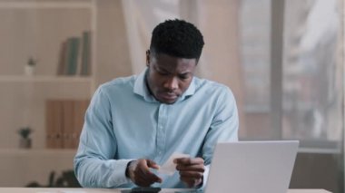 Genç Afrikalı adam odaklı banka muhasebecisi ödeme faturasını tutuyor kişisel bütçeyi yönetiyor hesap makinesi hesap makinası harcamaları hesaplıyor kredi bakiyesi ev ofis masasında oturarak hesaplama yapmaya devam ediyor