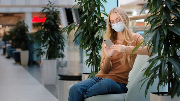 Donna caucasica impegnata in maschera medica seduta sul divano business a chiacchierare. Mascherato ragazza male centro commerciale femminile seduto sul divano guardando telefono cellulare acquisto online app navigazione messaggio di testo netto — Foto Stock