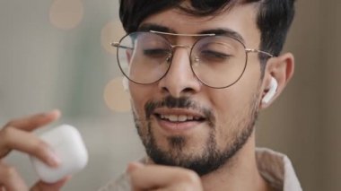 Yakın plan erkek mutlu yüz portresi Arap, İspanyol, sakallı, gözlüklü, kablosuz kulaklıklı, müzik dinleyen, modern kulaklıktaki sesin tadını çıkaran favori şarkısını söyleyen adam.
